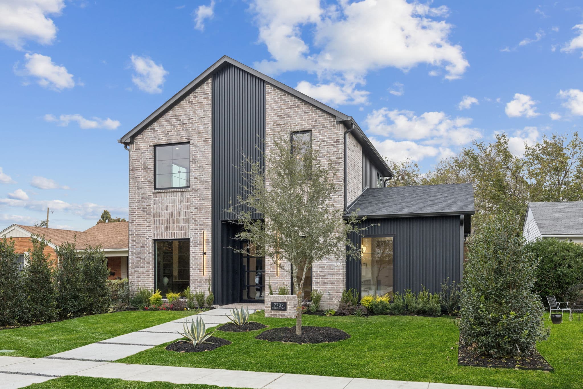 The front of 7707 Robin Road, a brick and dark metal house behind a young olive tree.